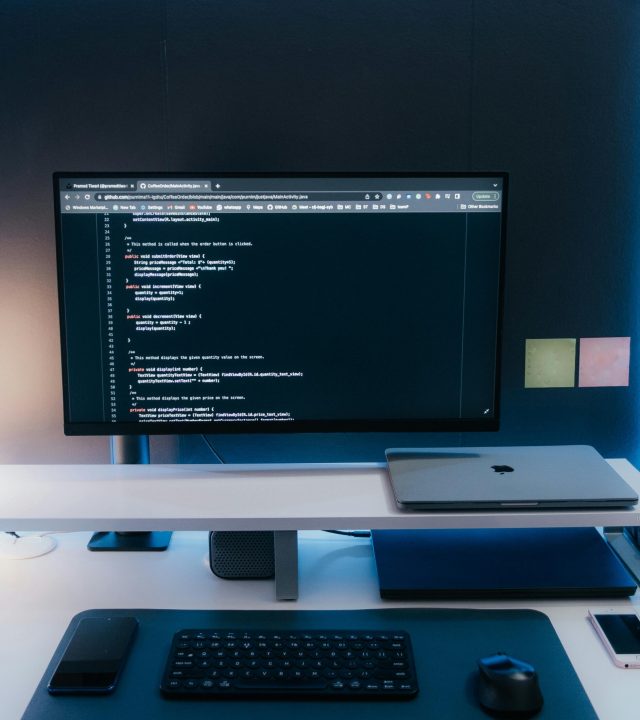 Minimalist home office desk setup with coding on a large monitor, laptop, and gadgets.